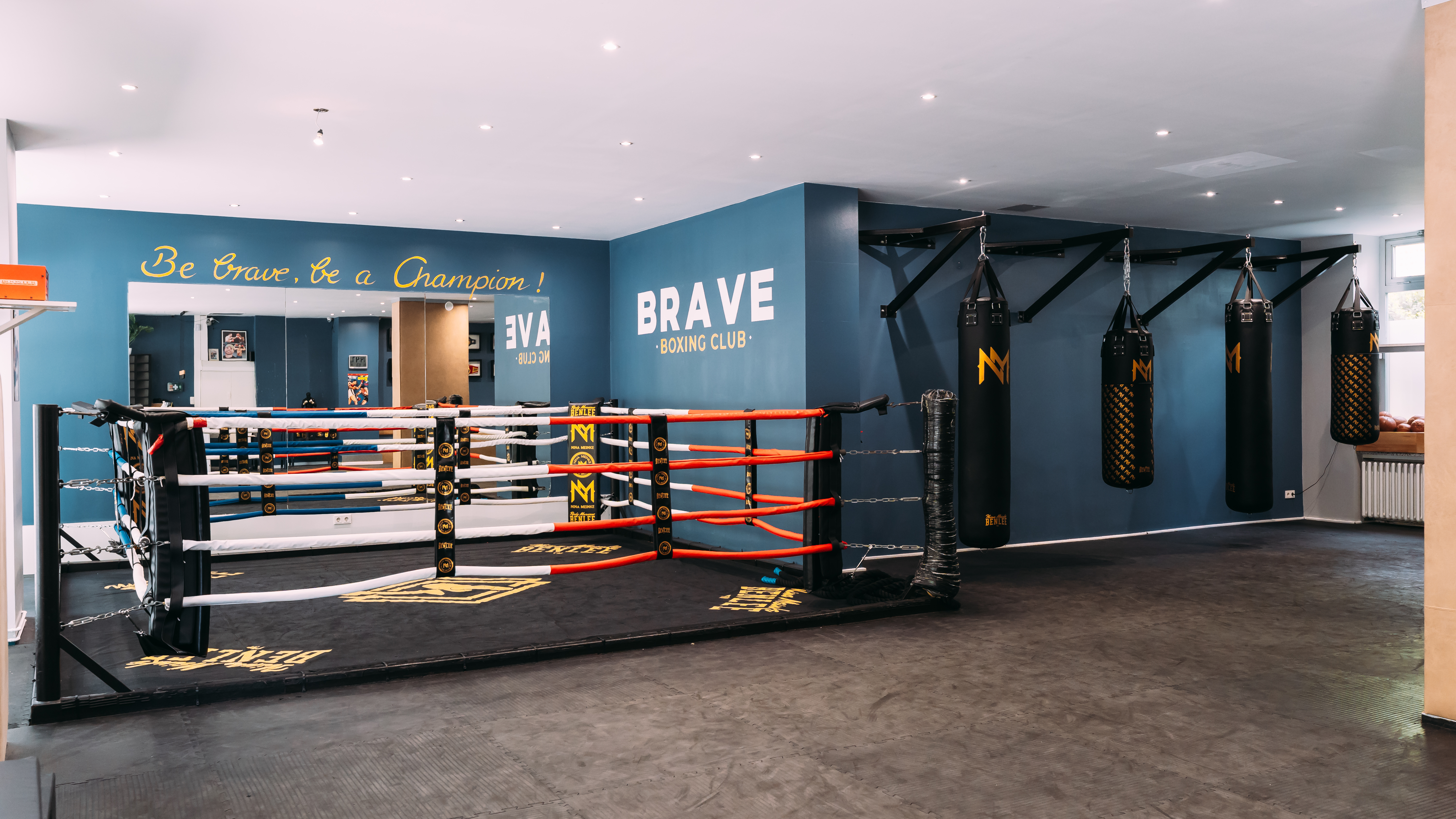 Im BRAVE BOXING CLUB stehen ein Boxring, mehrere schwere Sandsäcke und die Wandaufschrift „Be Brave, Be a Champion!“ in einem blauen Trainingsraum.