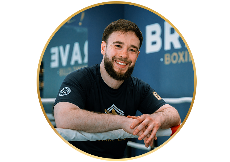 Lächelnder bärtiger Trainer lehnt im Boxring des Brave Boxing Club auf den Seilen und trägt ein schwarzes T-Shirt.