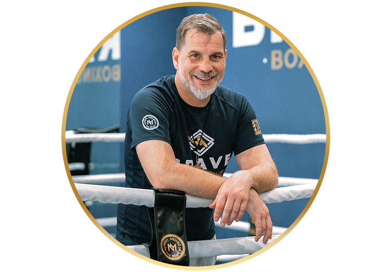 Ein lächelnder Boxtrainer lehnt im Ring an den Seilen und trägt ein schwarzes Shirt von The Brave Boxing Club.
