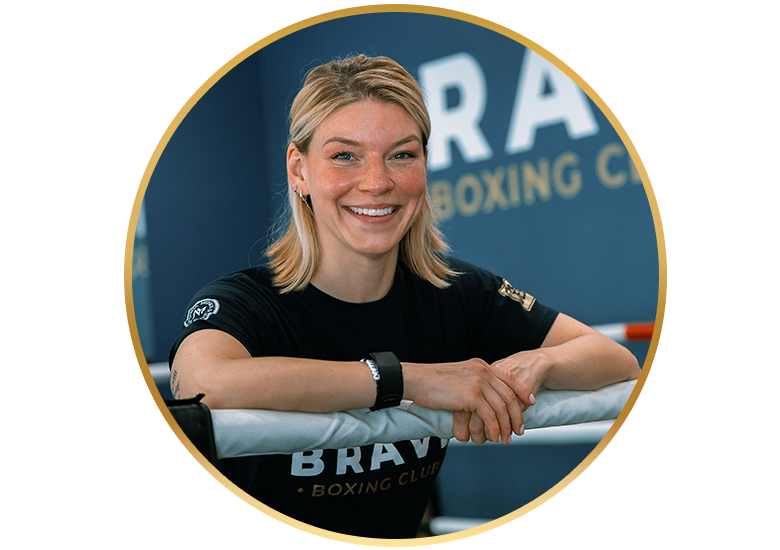Eine lächelnde Boxtrainerin lehnt im Ring an den Seilen, hinter ihr ist das Logo von The Brave Boxing Club zu sehen.
