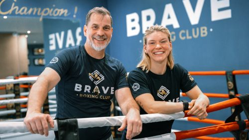 Zwei lächelnde Teammitglieder von The Brave Boxing Club lehnen an den Ringseilen vor einer blauen Wand.