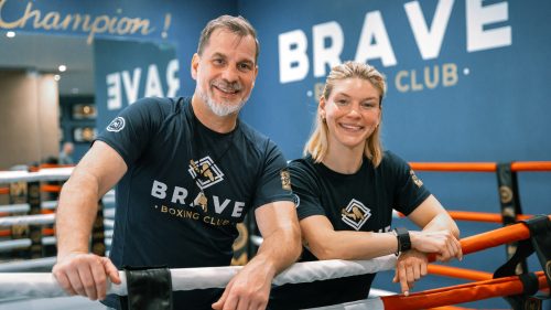 Ein Trainer und eine Athletin stehen lächelnd im Boxring des Brave Boxing Club und tragen schwarze Vereinskleidung.