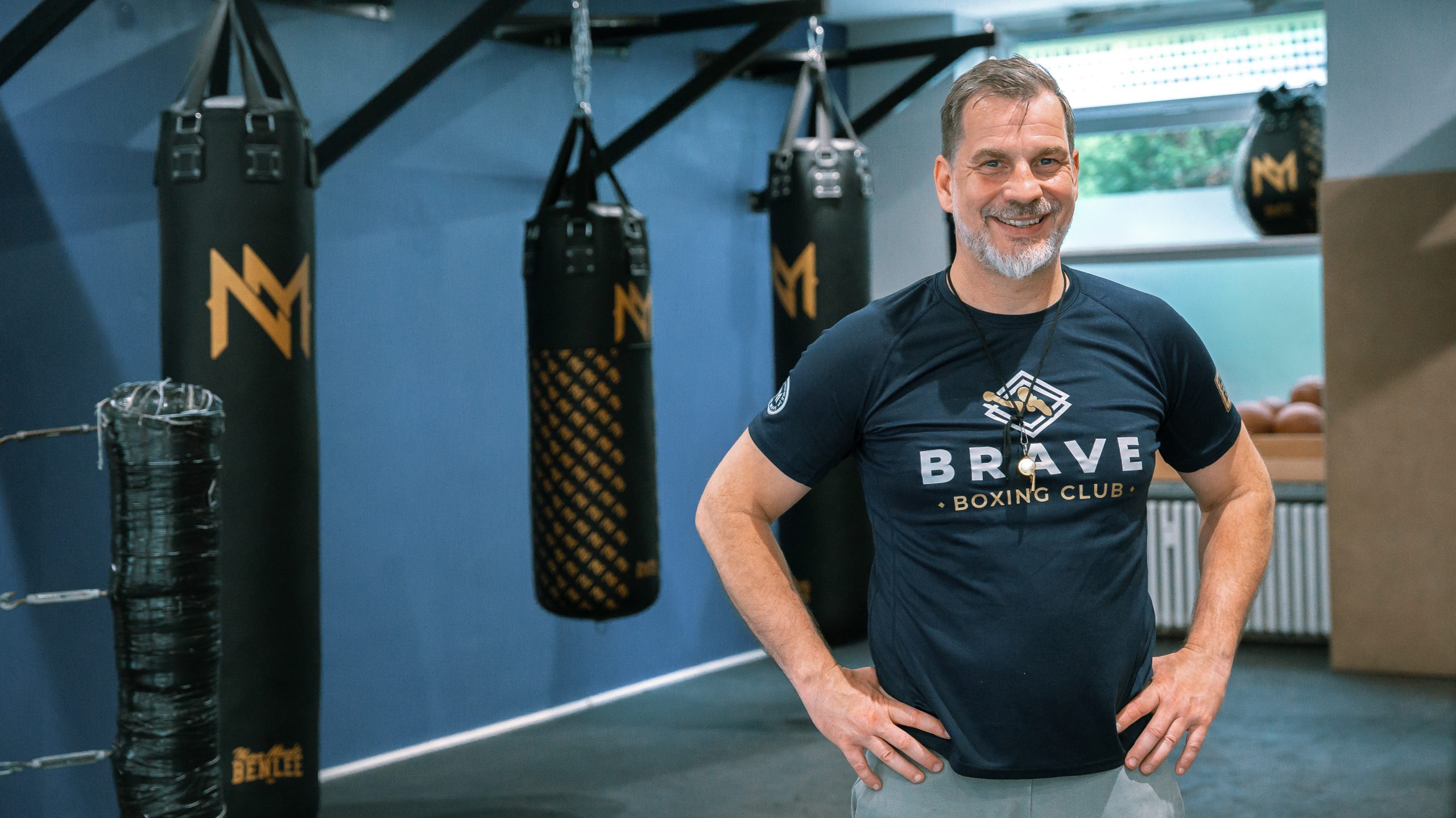 Ein lächelnder Mann steht im BRAVE Boxing Club vor hängenden Sandsäcken und trägt ein dunkles Shirt mit Vereinslogo.