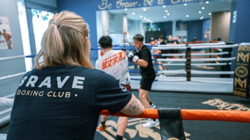 Im Brave Boxing Club beobachtet eine Frau mit blondem Zopf das Sparring im Ring, während zwei Boxer trainieren.