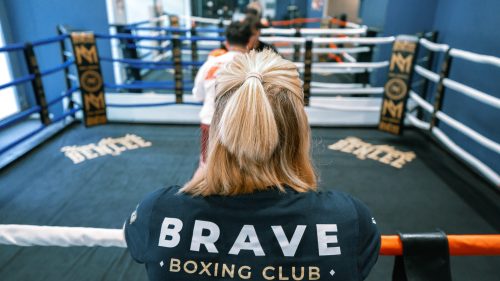 Eine blonde Person mit Pferdeschwanz steht im Boxring des Brave Boxing Club, während vor ihr eine weitere Person trainiert.
