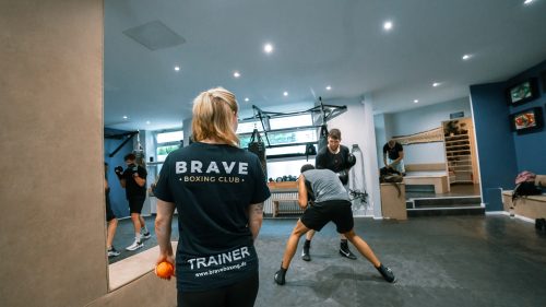 Im Brave Boxing Club leitet eine Trainerin mit blondem Haar eine Sparringseinheit, während mehrere Boxer im Gym trainieren.