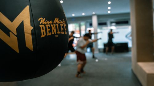 Ein schwarzer Benlee-Boxsack mit goldener Meinke-Schrift steht im Vordergrund, während Sportler im Hintergrund trainieren.