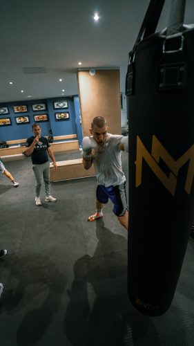 Ein Boxer mit weißen Handschuhen schlägt auf einen schwarzen Boxsack, während ein Trainer im Hintergrund zusieht.