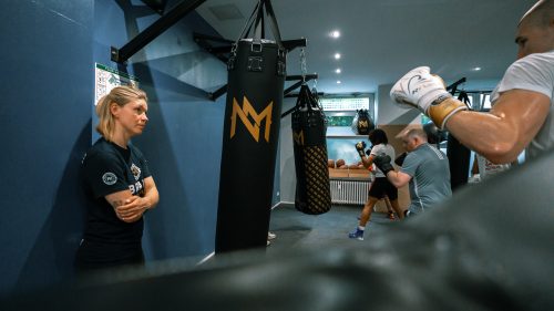 Im Meinke Gym beobachtet eine Trainerin das Boxtraining, während mehrere Sportler an Sandsäcken und im Sparring üben.