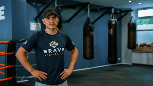 Ein Boxtrainer mit dunklem T-Shirt und schwarzem Cap steht im Brave Boxing Club vor mehreren Sandsäcken.