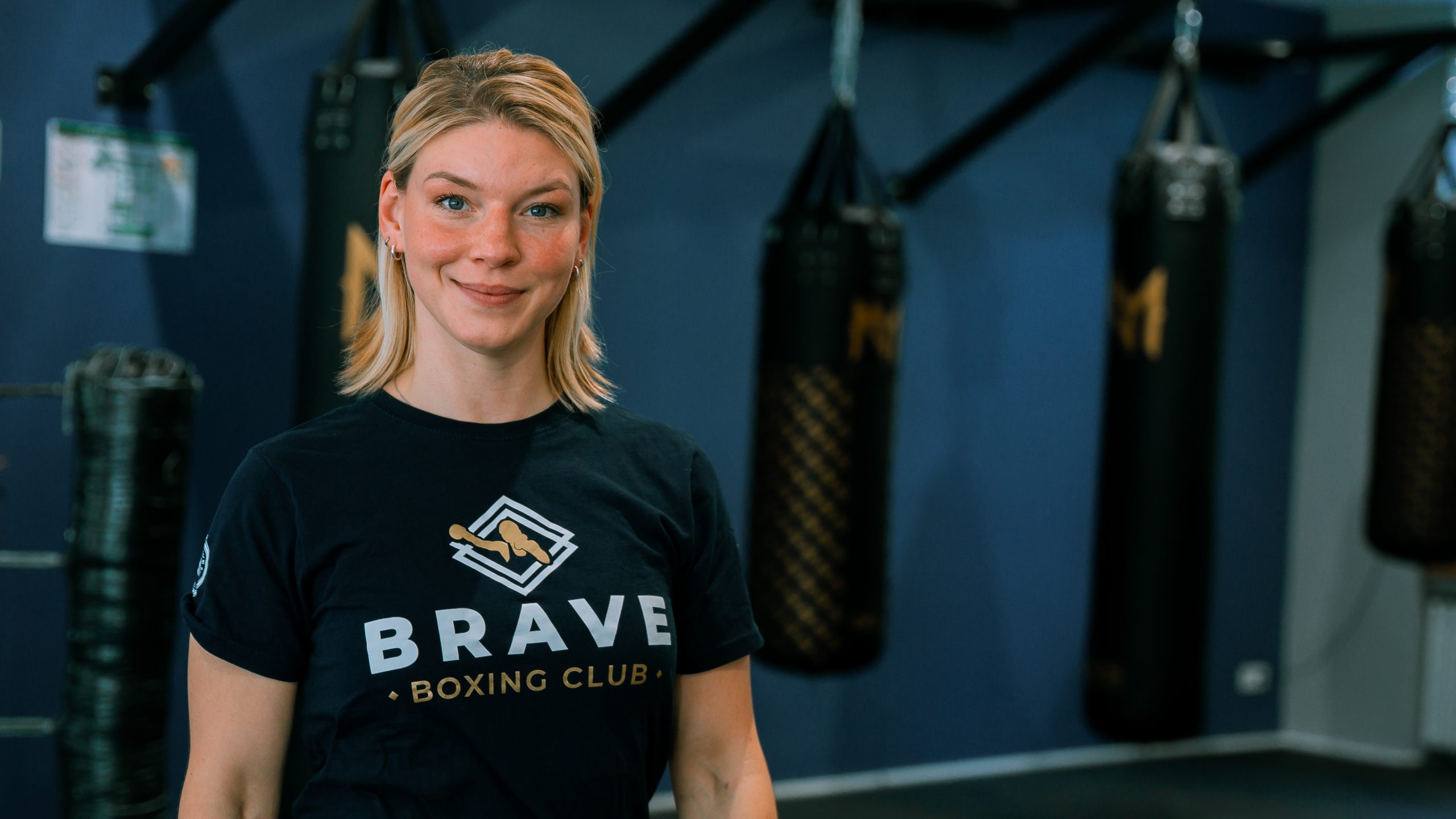 Eine lächelnde Boxtrainerin trägt ein Brave-Boxing-Club-Shirt vor schwarzen Boxsäcken im Trainingsraum.