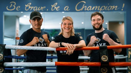 Drei lächelnde Boxerinnen und Boxer stehen im Ring des Brave Boxing Clubs und zeigen selbstbewusst ihre Fäuste.